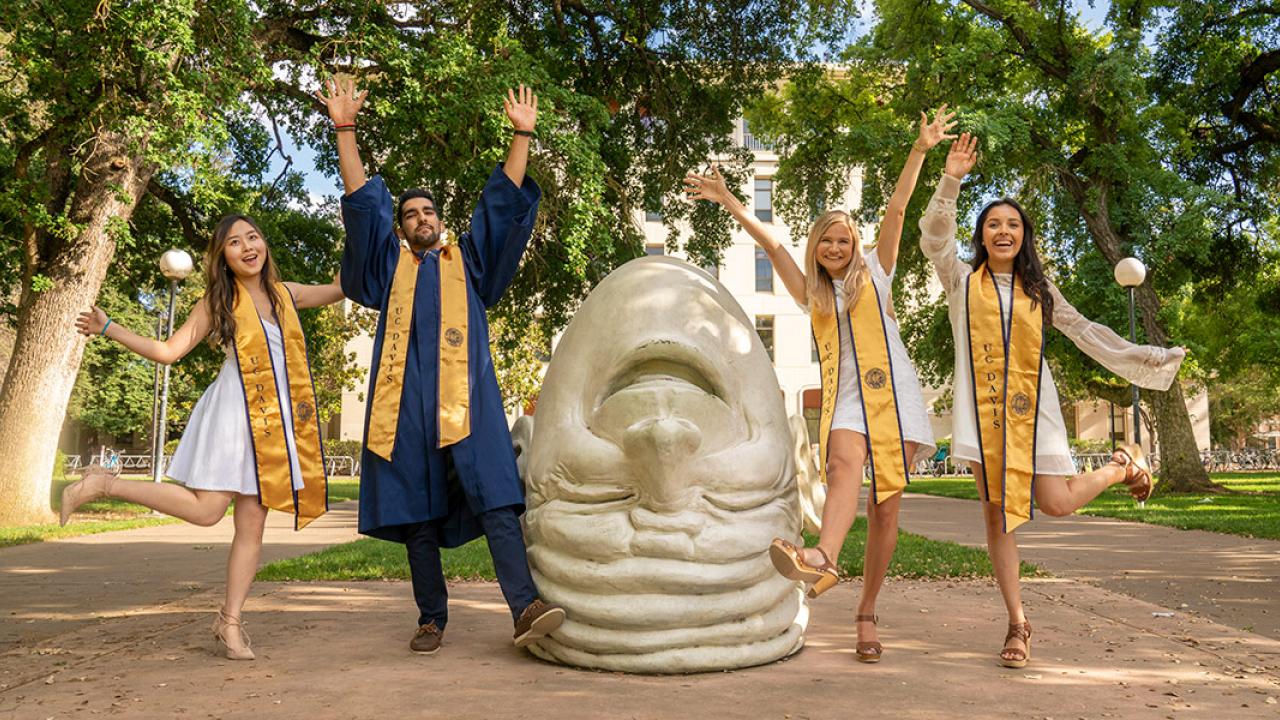 four students leaping up next to an Egghead sculpture, 'Eye on Mrak' in front of Mrak Hall
