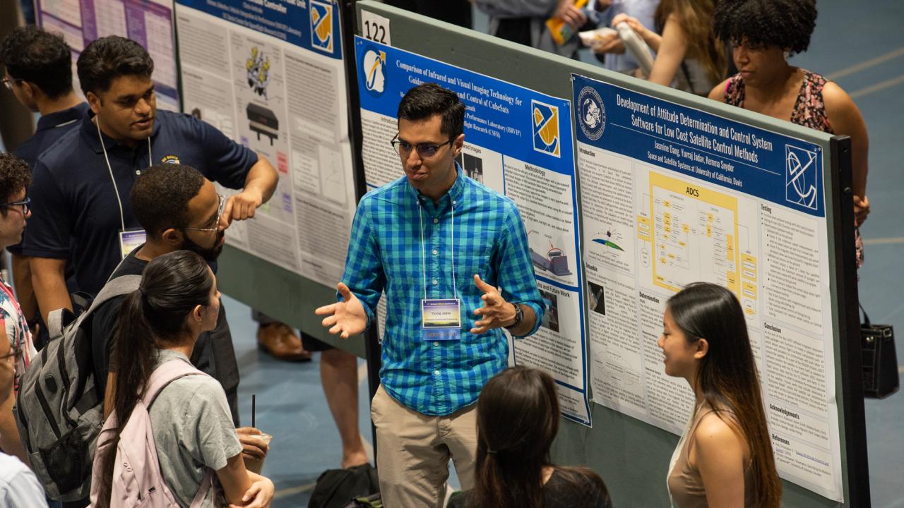 Student in blue shirt explains research to a half-circle of people gathered round poster display