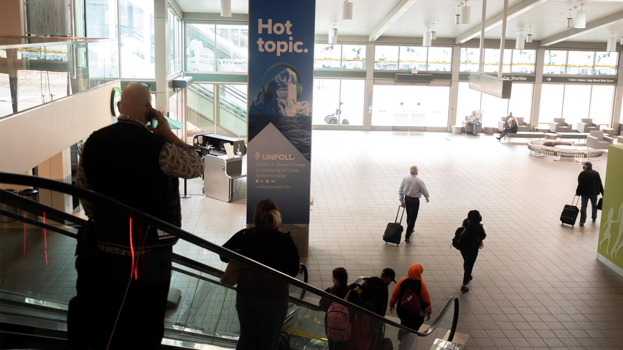 People descend an escalator at the Sacramento International Airport next to an ad for UC Davis’ Unfold podcast.