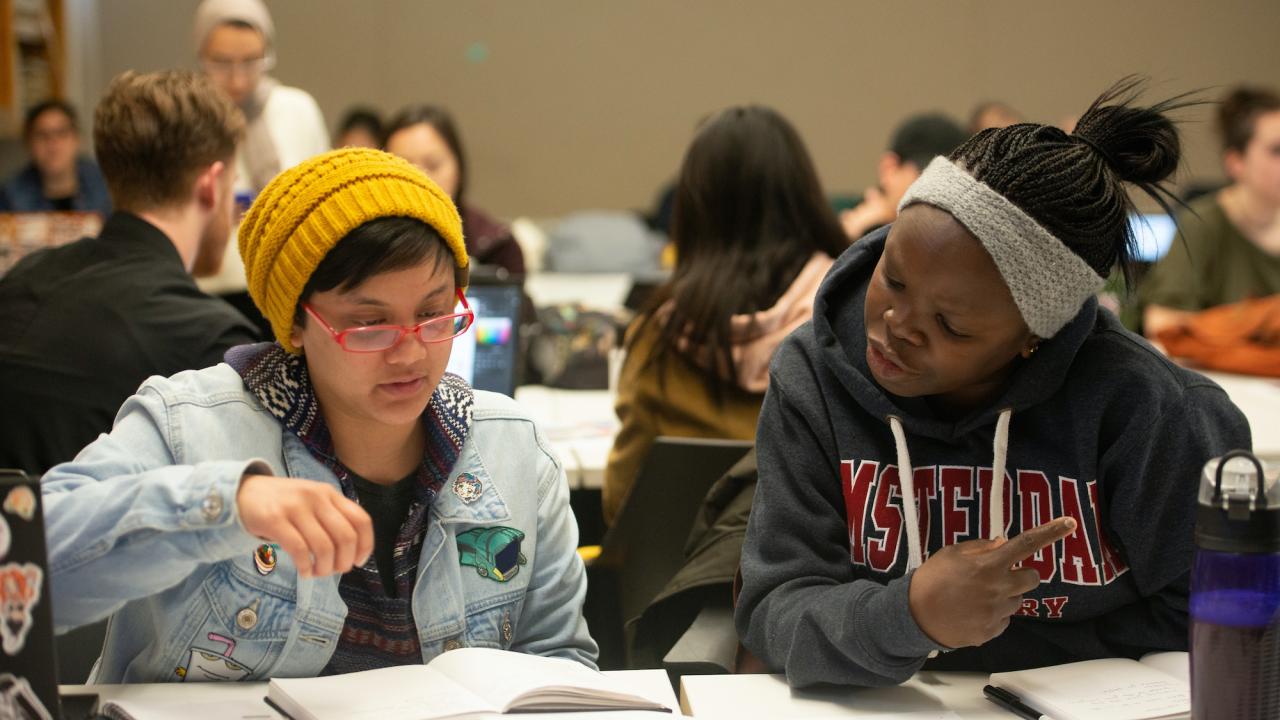 Two students look at notebooks and paper in a crowded classroom at UC Davis. 
