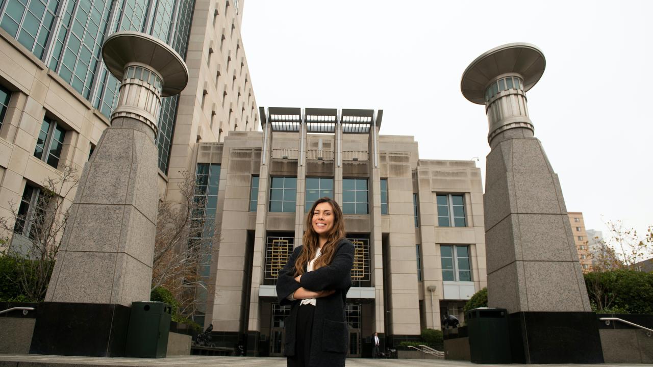 A political science major in a suit stands in front of a courthouse with her arms crossed