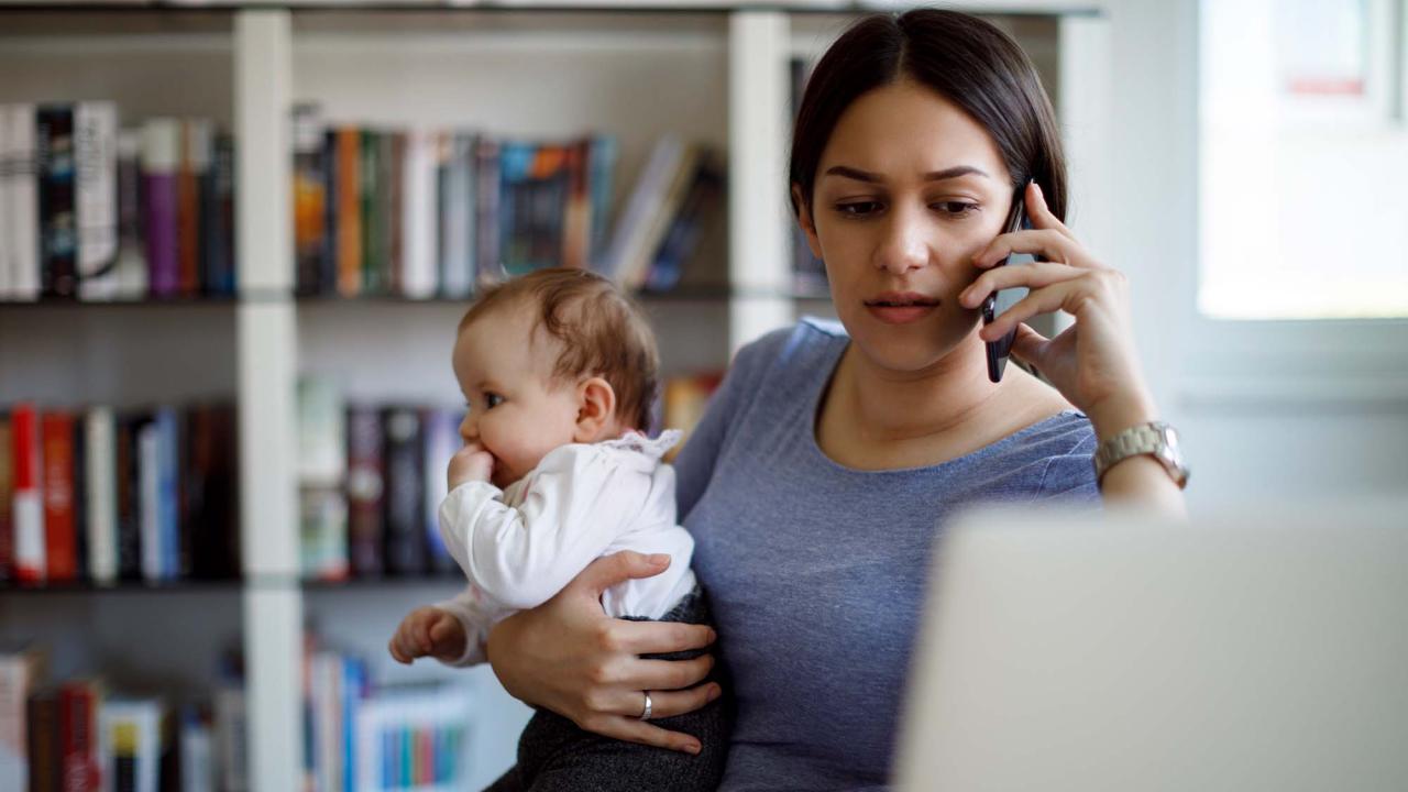 Woman holding small child while talking on phone. They sit at desk in front of a laptop screen.