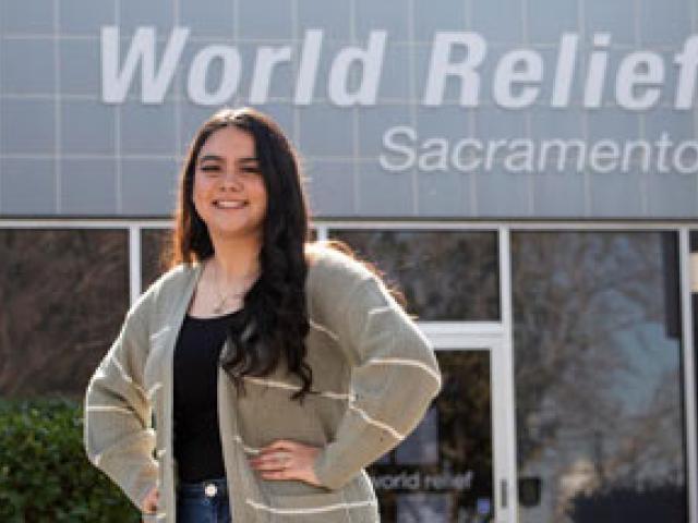 UC Davis junior Cristina Lopez, posing, “World Relief Sacramento” sign on building behind her