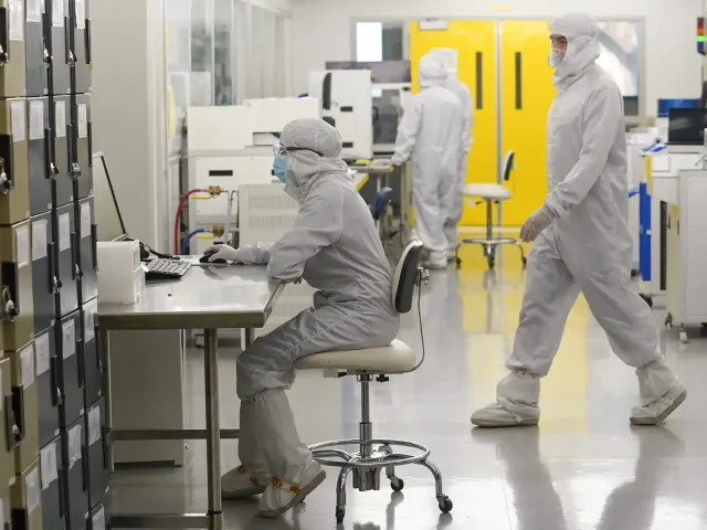 Researchers wearing full cleanroom suits work inside a bright, sterile laboratory. One person sits at a metal workstation using a computer, while others walk or operate equipment in the background. Rows of storage lockers line one side of the room, and large yellow doors are visible at the back.