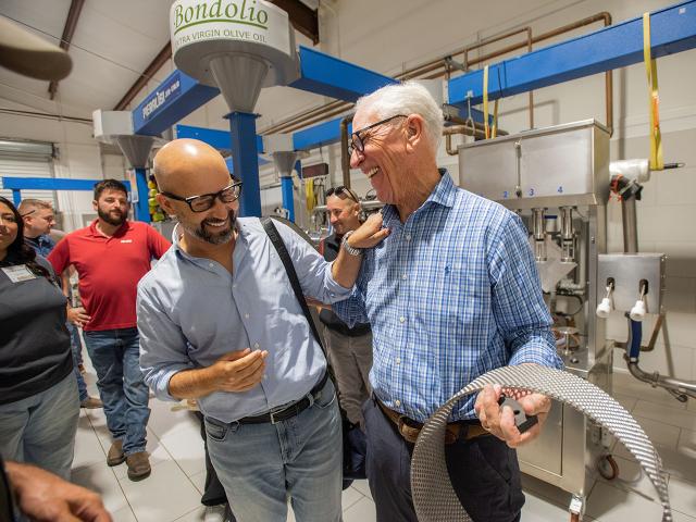Two men talking in a lab; older man holds coiled tubing among machinery