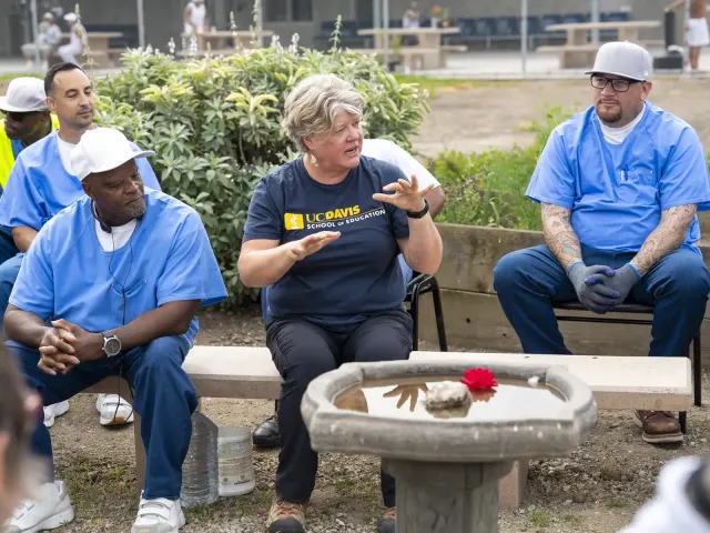 Group seated outdoors around a low planter; a woman gestures while others listen.