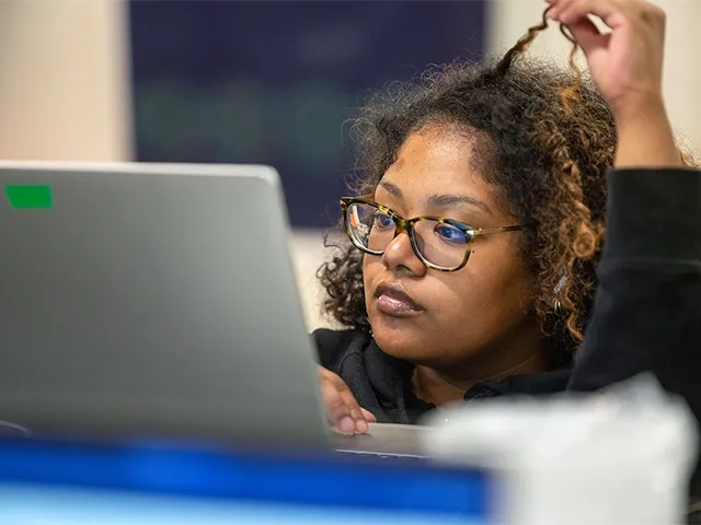 Student with glasses focused on laptop, twirling curly hair.