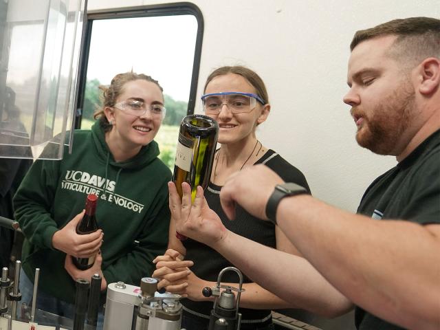 A Halsey Bottling employee holds up a wine bottle and points to it while explaining a point to two smiling students in safety goggles.