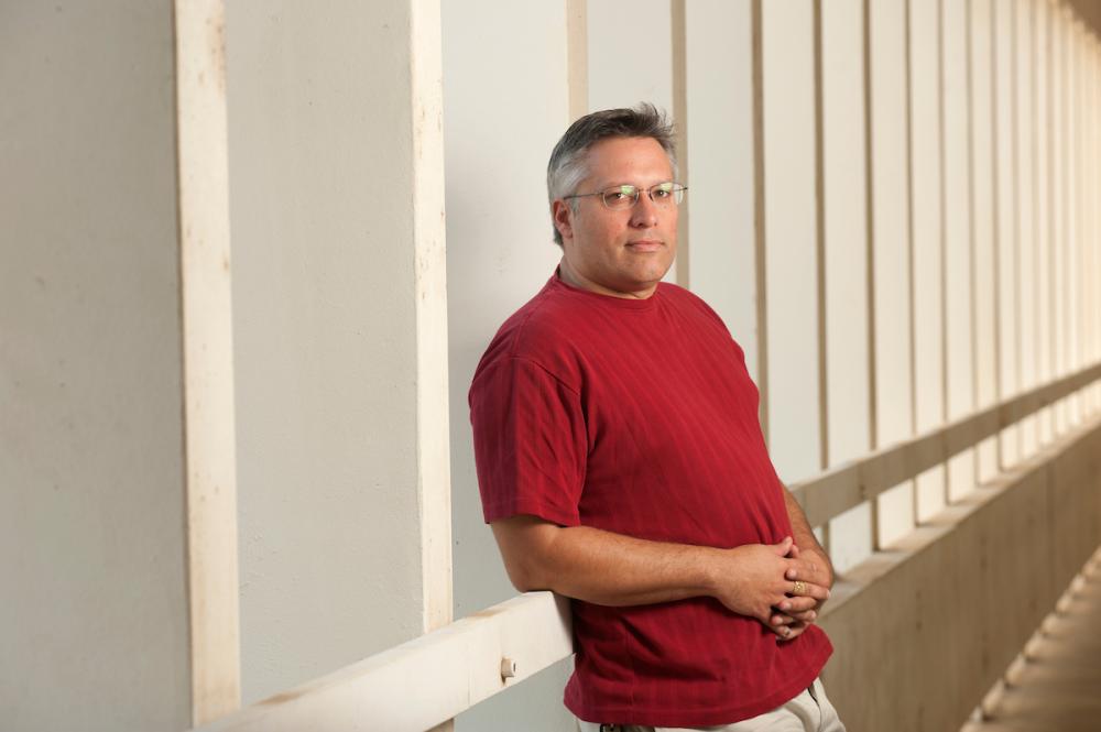 Grey-haired man in red t-shirt leans against a breezeway wall. 
