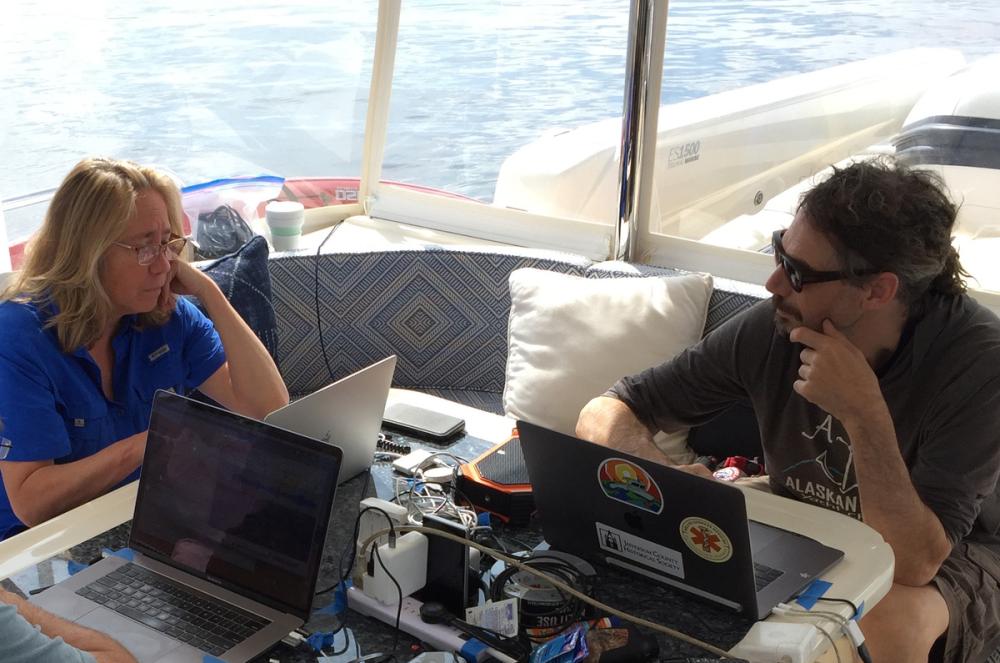 A blonde woman in blue shirt and glasses and a man with dark curly hair and dark glasses work on laptops aboard a boat. 