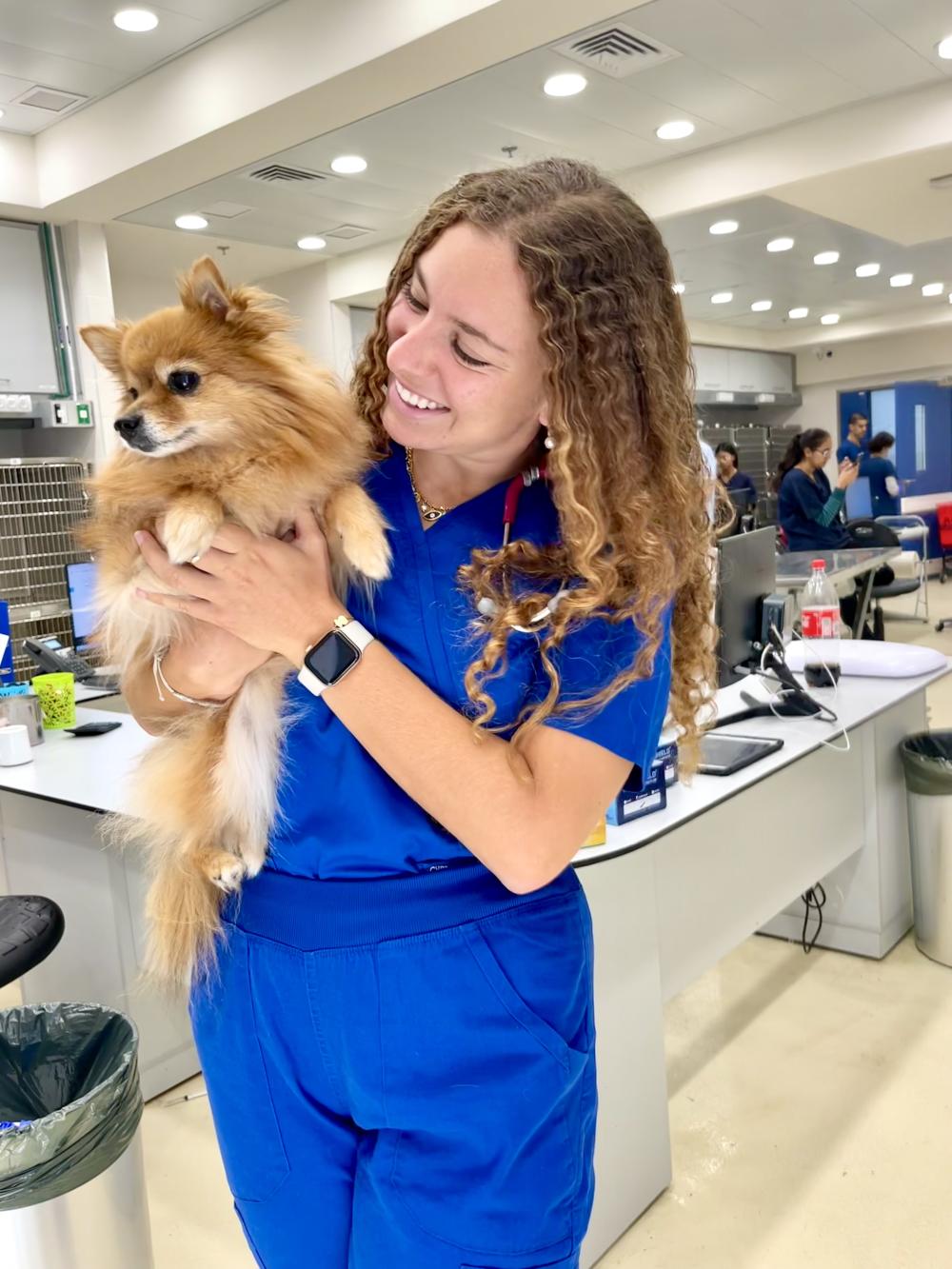 Orli Algrannati, current animal science student, wears her blue scrubs while holding a fluffy light brown dog.