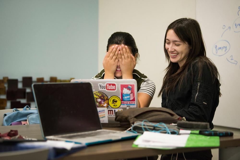 Two college students laugh as they look at a laptop computer.
