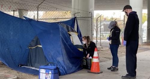 Three people stand outside of a tent under a freeway overpass
