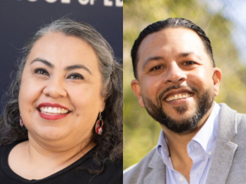 A woman with curly hair and earrings smiles alongside a man in a grey blazer, both cheerful.