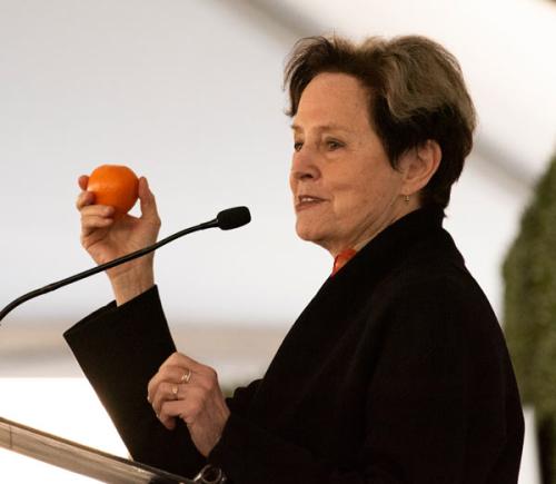 Woman at lectern, holding a piece of fruit
