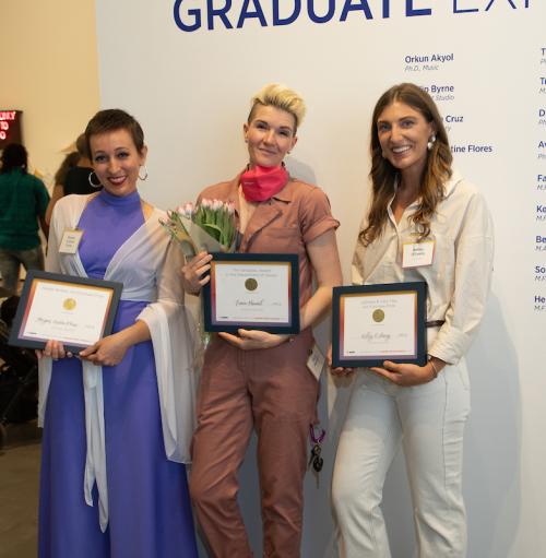 Three award-winning artists hold their certificates as they stand in front of exhibit wall.