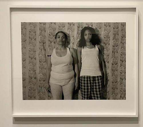 A black and white photo by LaToya Ruby Frazier depicts her and her mother standing side by side against a patterned wall. Both women are dressed in their pajamas and stare directly at the camera.