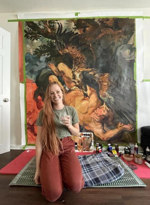 A woman kneels and smiles in front of a floor to ceiling painting of a large bird attacking a man.