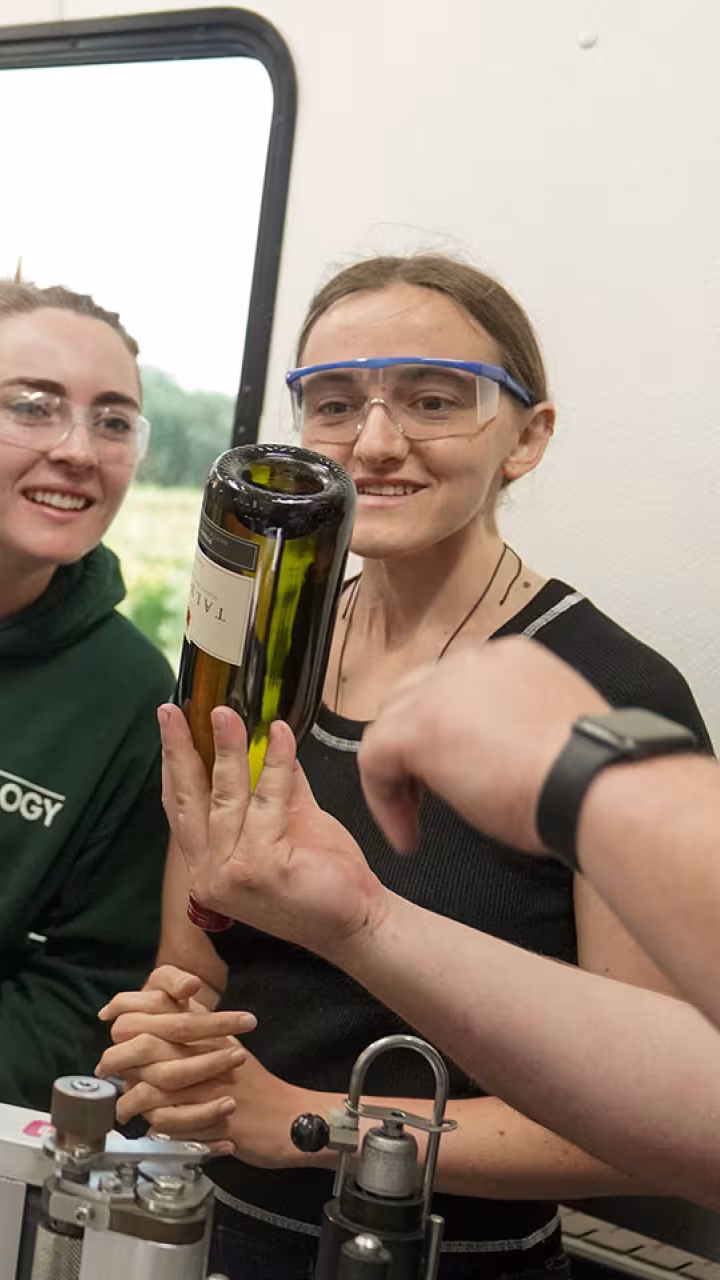 A Halsey Bottling employee holds up a wine bottle and points to it while explaining a point to two smiling students in safety goggles.