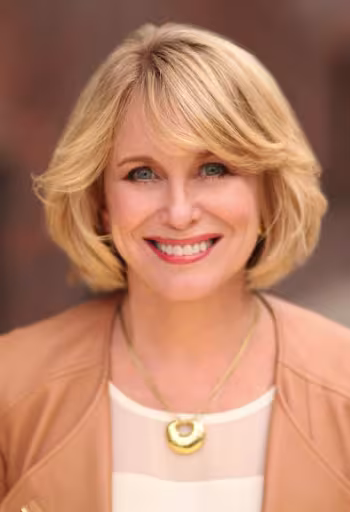 Smiling woman with blonde hair, wearing a beige jacket and gold necklace, outdoors.