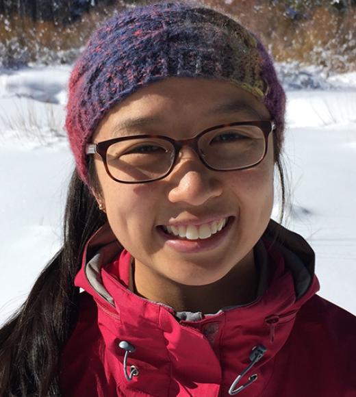 Head and shoulders of a smiling Asian woman wearing glasses, a red ski jacket and purple beanie. 