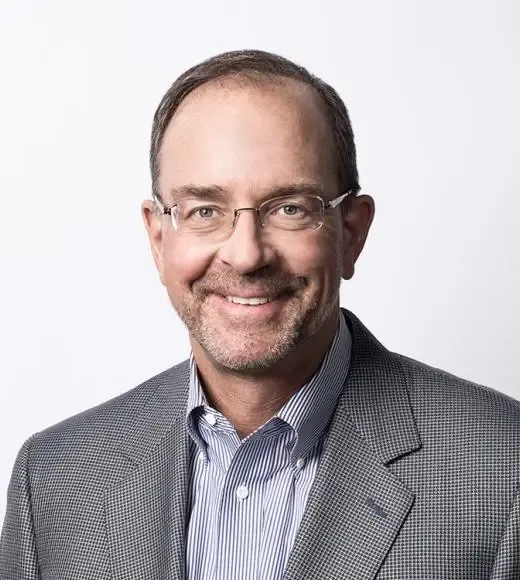 Smiling man with glasses, wearing a gray blazer and blue shirt, against a plain background.