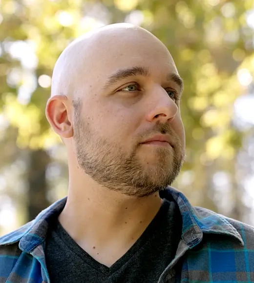 A bald man with a beard stands outdoors, wearing a plaid shirt, looking thoughtfully to the side.