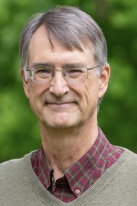 Smiling older man with glasses outdoors, green sweater over red plaid shirt