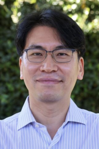 Smiling man wearing glasses and a striped collared shirt, blurred green foliage background