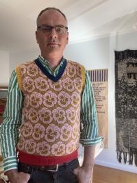 Man wearing glasses and pink floral sweater vest over green striped shirt, by wall tapestry