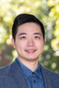 Smiling man wearing a blue shirt and blazer, with a blurred outdoor background.