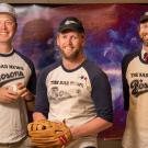 Three men, softball players, in "The Bad News Bosons" jerseys. One man holds a softball, another has a glove on.