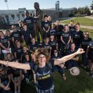 Janice Corbett, arms outstretched, with Warriors behind her, around Jim Sochor statue outside Aggie Stadium