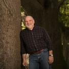 Man in jeans and plaid shirt stands next to trunk of large tree