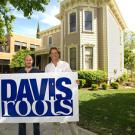 Two men standing behind a Davis Roots sign with a old mansion in the background