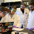 Four students wearing lab coats, goggles and gloves stand near a laptop.