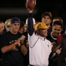 Coach Fred Aro holds football in air, as if in triumph, on sidelines.