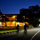 Photo: Two bicyclists on path approachining well-lit Wellman Hall at night.l