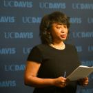 Woman reads from script against "UC Davis" backdrop.