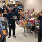Mail Services crew and UC Davis Health executive hold food items, while standing around tables filled with food donations.