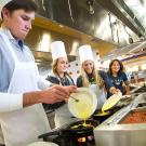 Photo: Students pour eggs into scrambler dishes, at a stove in the dining commons.