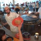 Photo: chef hands bowl of noodle to woman, with many people in the background