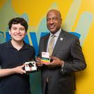 A young man in a black polo shirt holds a Polaroid camera made out of Legos against his chest. He is standing to the left next to Chancellor Gary S. May who is wearing a dark suit and holding a small box of Polaroid film. Both are smiling at the camera in a studio setting with a bright background that has blue UC Davis logo along a yellow wall.