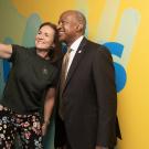 A woman and a man smiling together while taking a selfie against a colorful background.