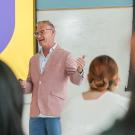 Man in pink blazer lecturing at a whiteboard to seated students