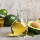 Glass bottle and bowl of golden avocado oil with whole and halved avocados on gray surface