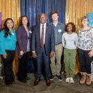 Eight adults standing side-by-side, smiling at indoor event with blue and gold backdrop