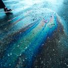 a rainbow sheen of oil on wet pavement and the legs of someone standing near it