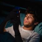 Young man in bed, looking at his phone in a dimly lit room.