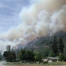 Smoke from wildfire billows over hillside with trees and rural home in foreground